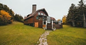 Rustic wooden cabin surrounded by autumn trees and lush greenery, featuring a stone path leading up to the entrance.